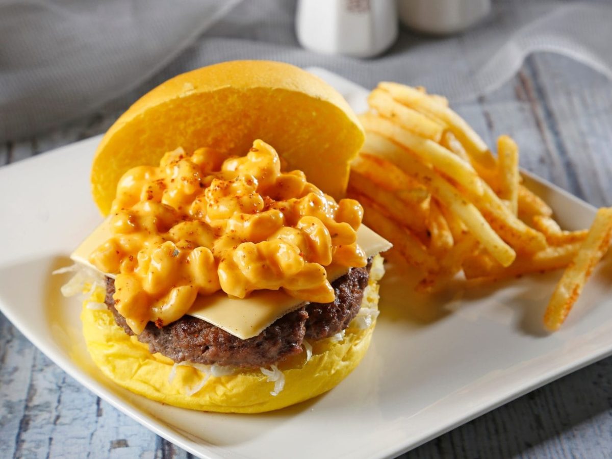 Mac'n Cheese Burger mit Coleslaw auf weißem Teller mit Pommes auf hellem Untergrund. Frontalansicht.