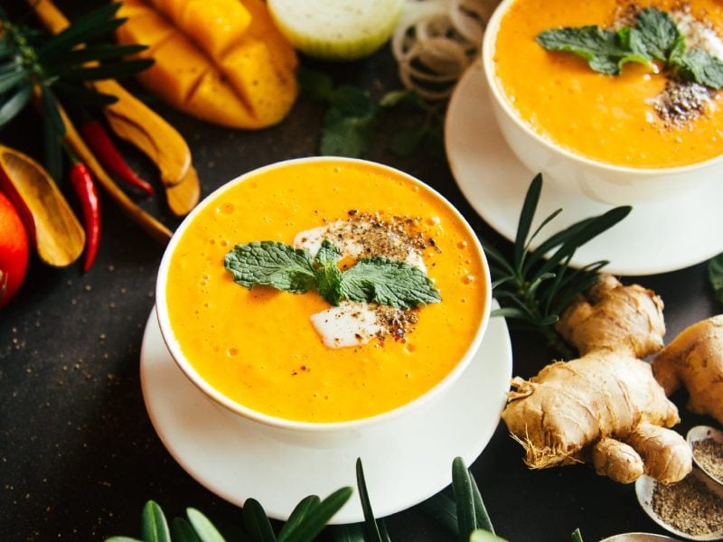 Zwei Suppenschalen mit der gelben Suppe auf dunklem Untergrund und allerlei Zutaten und Gewürzen daneben.