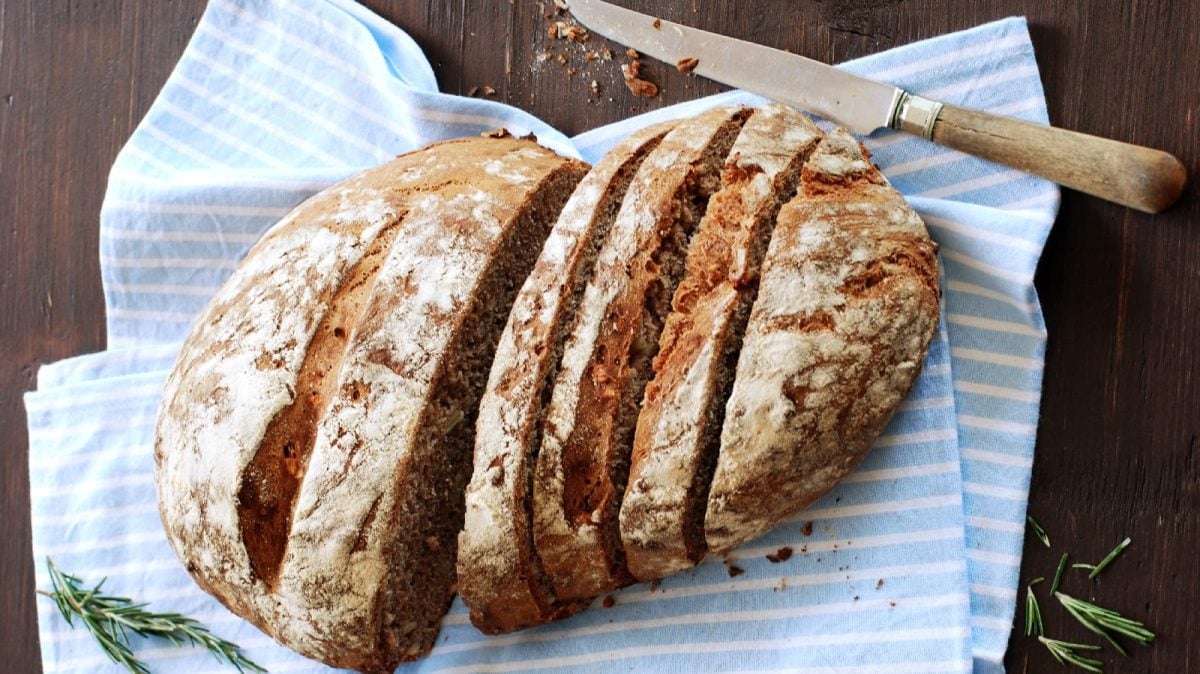 Draufsicht: Aufgeschnittenes rustikales Maronenbrot auf einem blau-weiß gestreiften Geschirrtuch. Liegend auf einer dunkelbraunen Tischplatte.