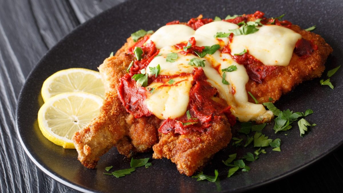 Milanesa Napolitana mit Zitrone und Petersilie auf schwarzem Teller und grauem Untergrund. Draufsicht.