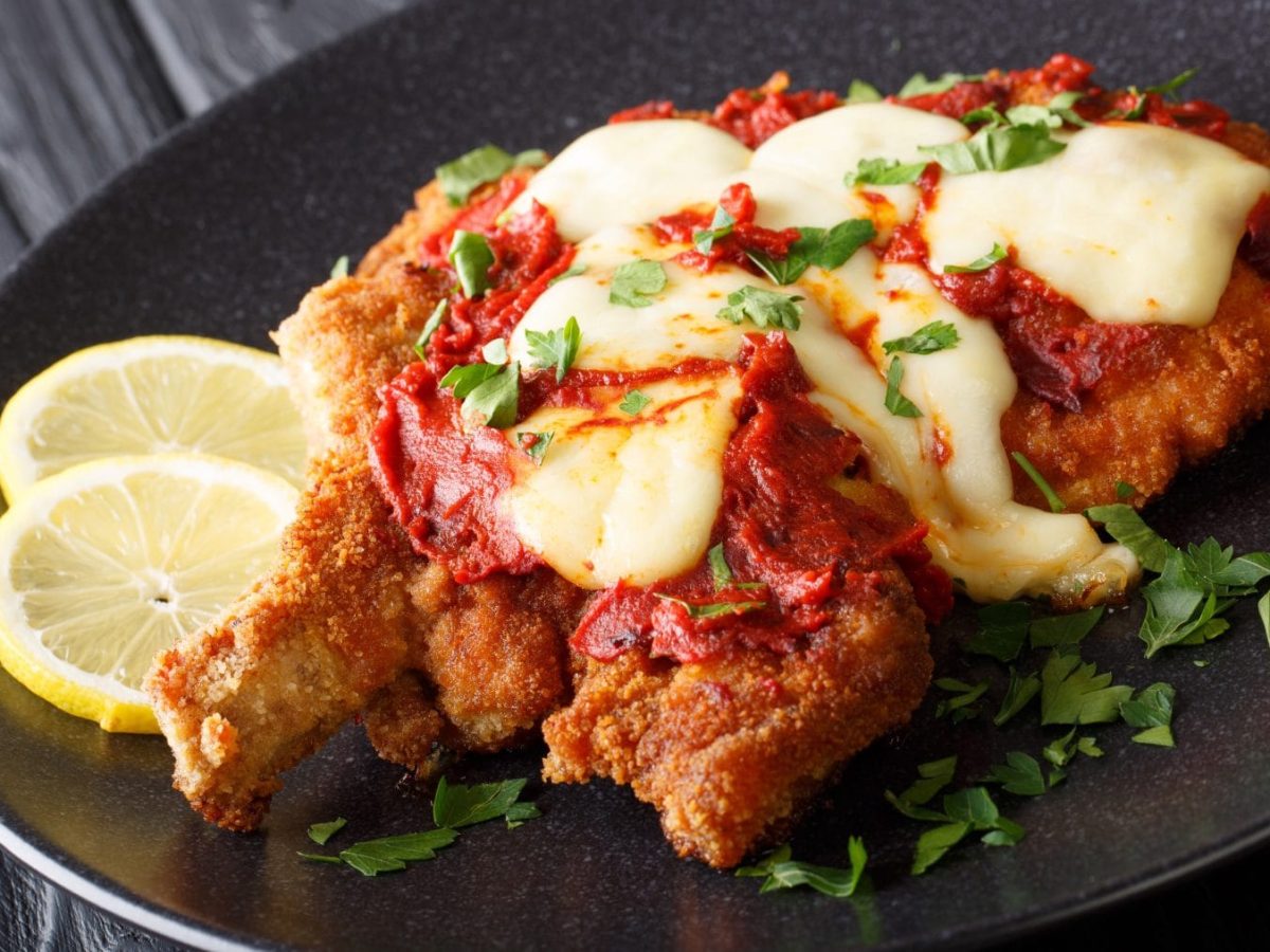 Milanesa Napolitana mit Zitrone und Petersilie auf schwarzem Teller und grauem Untergrund. Draufsicht.