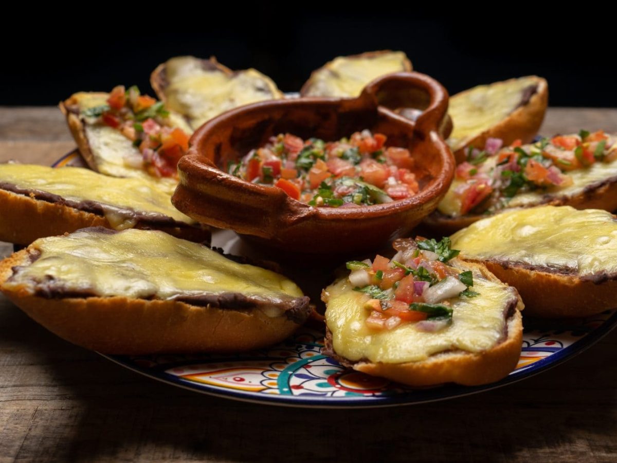 Molletes mit schwarzem Bohnenmus und Käse überbacken auf buntem Teller. In der Mitte eine Schale mit Pico de Gallo. Platziert auf Holz. Frontalansicht.