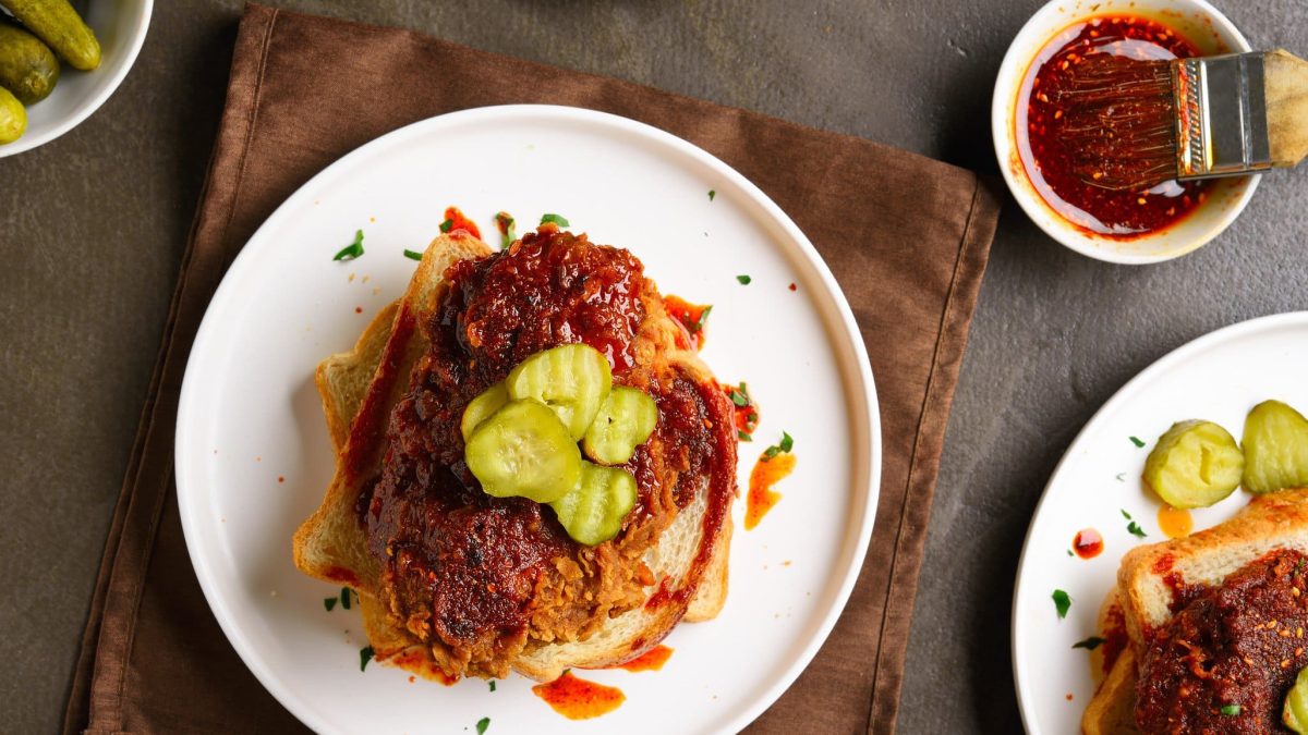 Nashville Hot Chicken aus weißem Teller auf braunem Tuch und Holztisch. Daneben eine Schale mit Chili-Öl und Pinseln. Draufsicht.