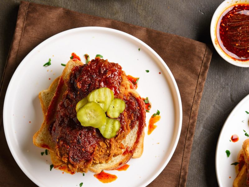 Nashville Hot Chicken aus weißem Teller auf braunem Tuch und Holztisch. Daneben eine Schale mit Chili-Öl und Pinseln. Draufsicht.