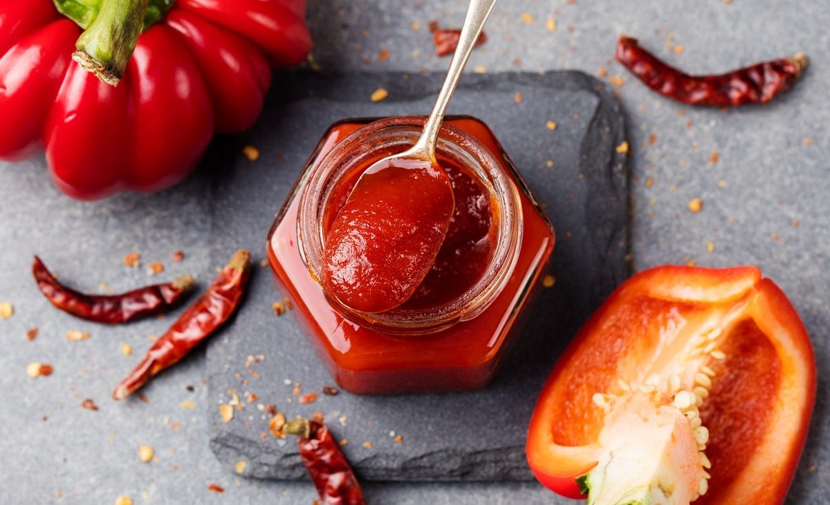 Draufsicht: In einem Glas ist eine Rote, cremige Masse. Auf dem Glas liegt ein teelöffel mit der Masse. Drum herum liegt eine ganze Paprika, eine halbe Paprika sowie Chilischoten.