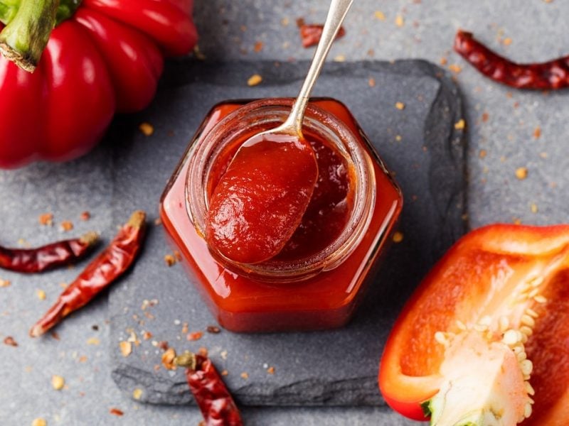 Draufsicht: In einem Glas ist eine Rote, cremige Masse. Auf dem Glas liegt ein teelöffel mit der Masse. Drum herum liegt eine ganze Paprika, eine halbe Paprika sowie Chilischoten.