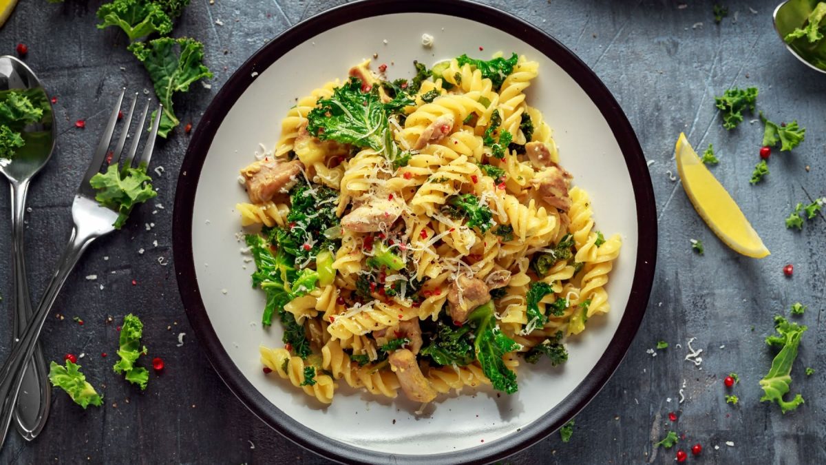 Ein weißer Teller mit schwarzem Rand auf grauem Untergrund. Darauf das PAstagericht mit Hähnchen und Grünkohl.