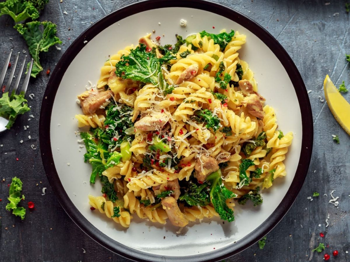 Ein weißer Teller mit schwarzem Rand auf grauem Untergrund. Darauf das PAstagericht mit Hähnchen und Grünkohl.