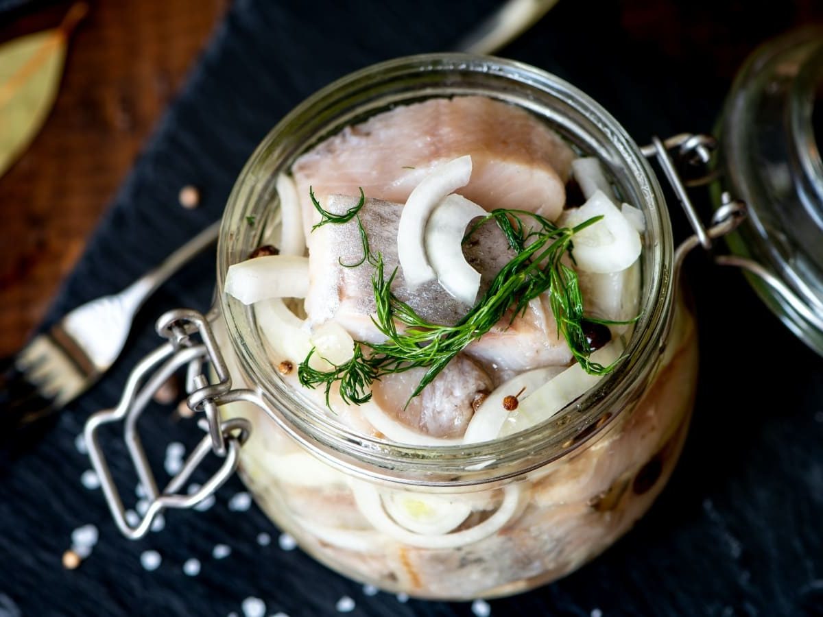 Heringstücke mit Zwiebelscheiben und Pfefferkörnern in einem Bügelglas auf schwarzem Untergrund.