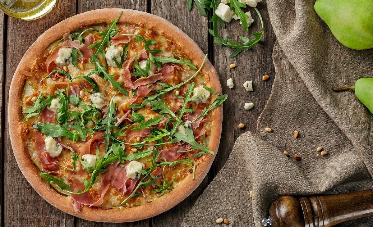 Draufsicht: Eine Pizza mit Schinken, Birne und Gorgonzola liegt auf einem Untergund aus Holz. Daneben liegen Birnen, Käsewürfel, Pinienkerne, eine Pfeffermühle und ein Tuch aus Leinen.