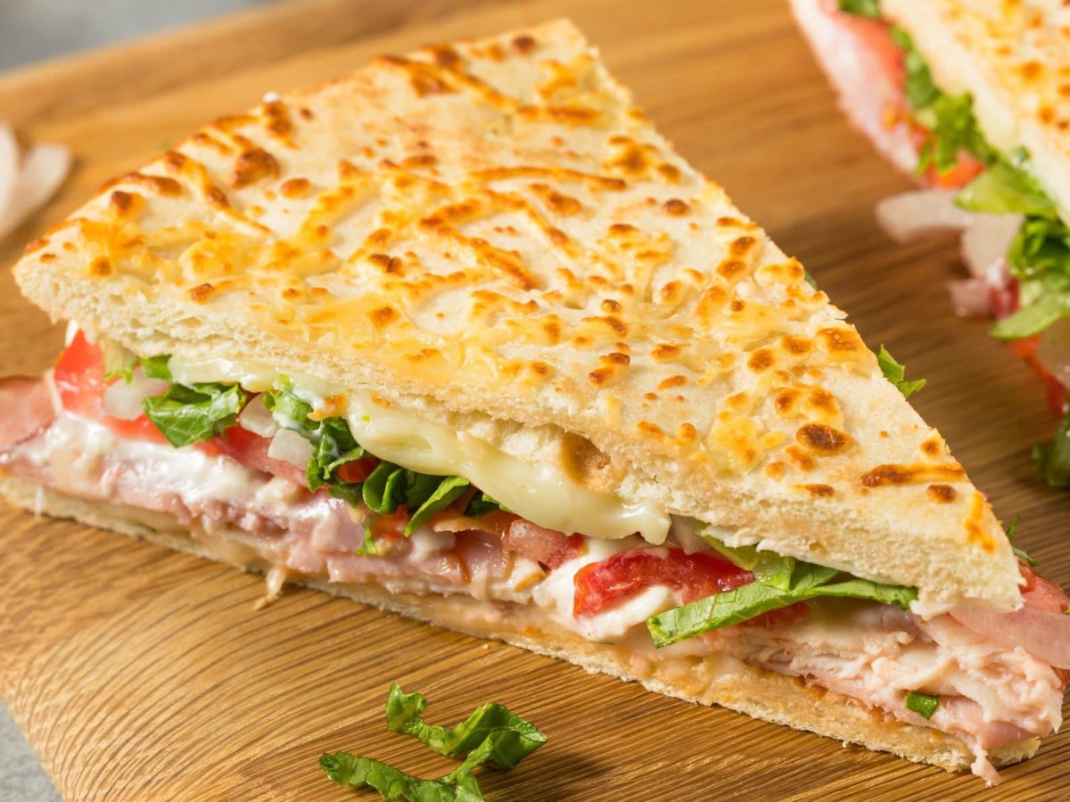 Ein Stück Pizza-Sandwich mit Schinken, Käse und Tomaten auf einem hellen Holzschneidebrett.