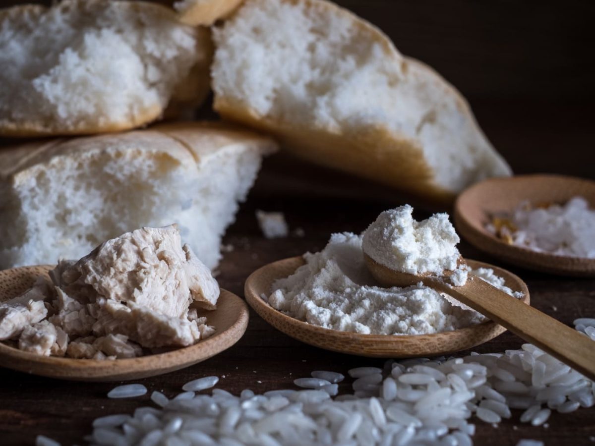 Das Reisbrot im Hintergrund. Im Vordergrund drei kleine Holzschippen mit Reismehl, Hefe und körnigem Reis.