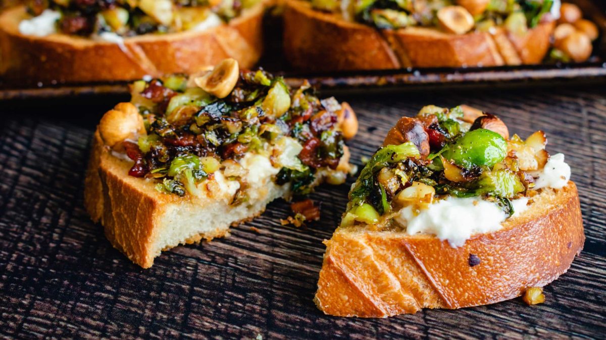 Mehre Scheiben Brot auf einem dunkelgrauem Untergrund mit Rosenkohl, Speck und Burrata.