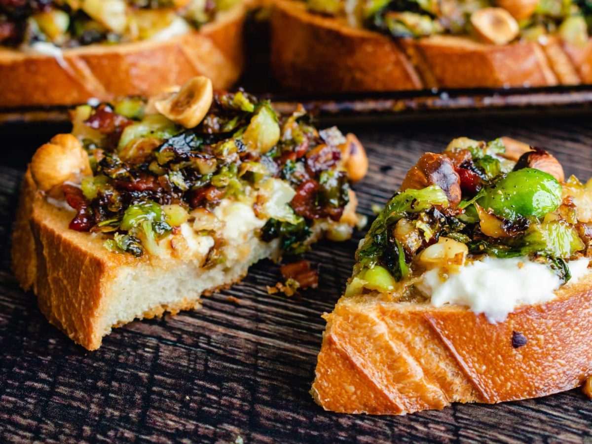 Mehre Scheiben Brot auf einem dunkelgrauem Untergrund mit Rosenkohl, Speck und Burrata.