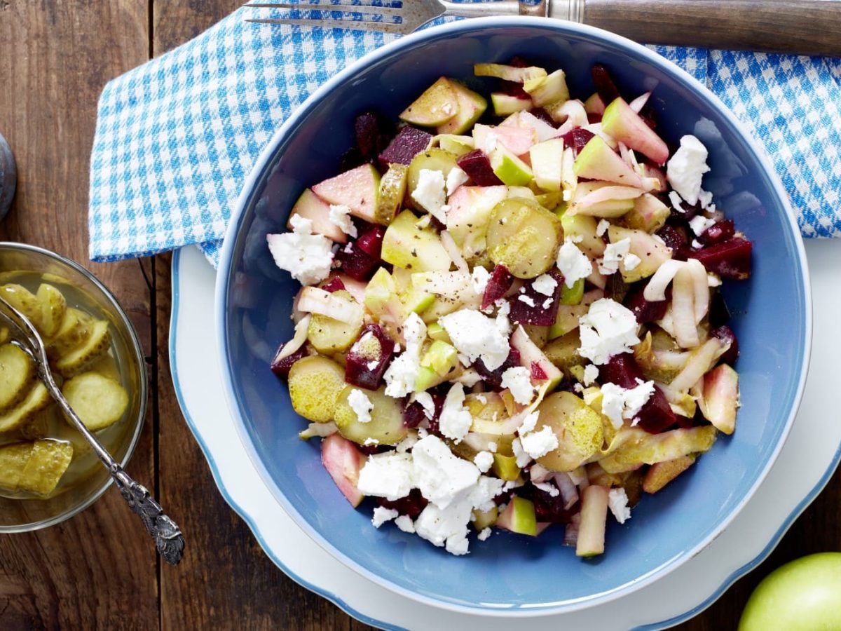 Rote-Bete-Salat mit Feta in blauer Schüssel, daneben Gewürzmühle, saure Gurken und ein grüner Apfel vor Holzhintergrund.