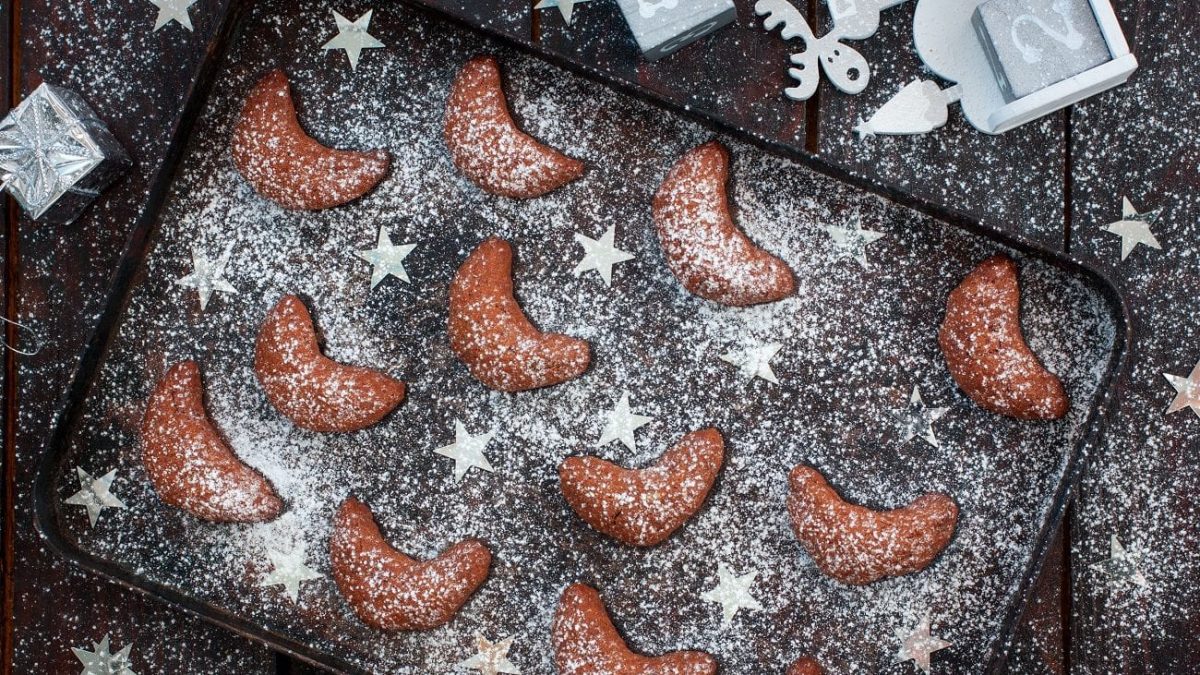 Draufsicht: Auf einem dunklen Backblech liegen dunkle schokoladen-kipferl. Die sind mit einem hellen Puder bestäubt. Dazwischen liegen Sterne aus hellem Metall.