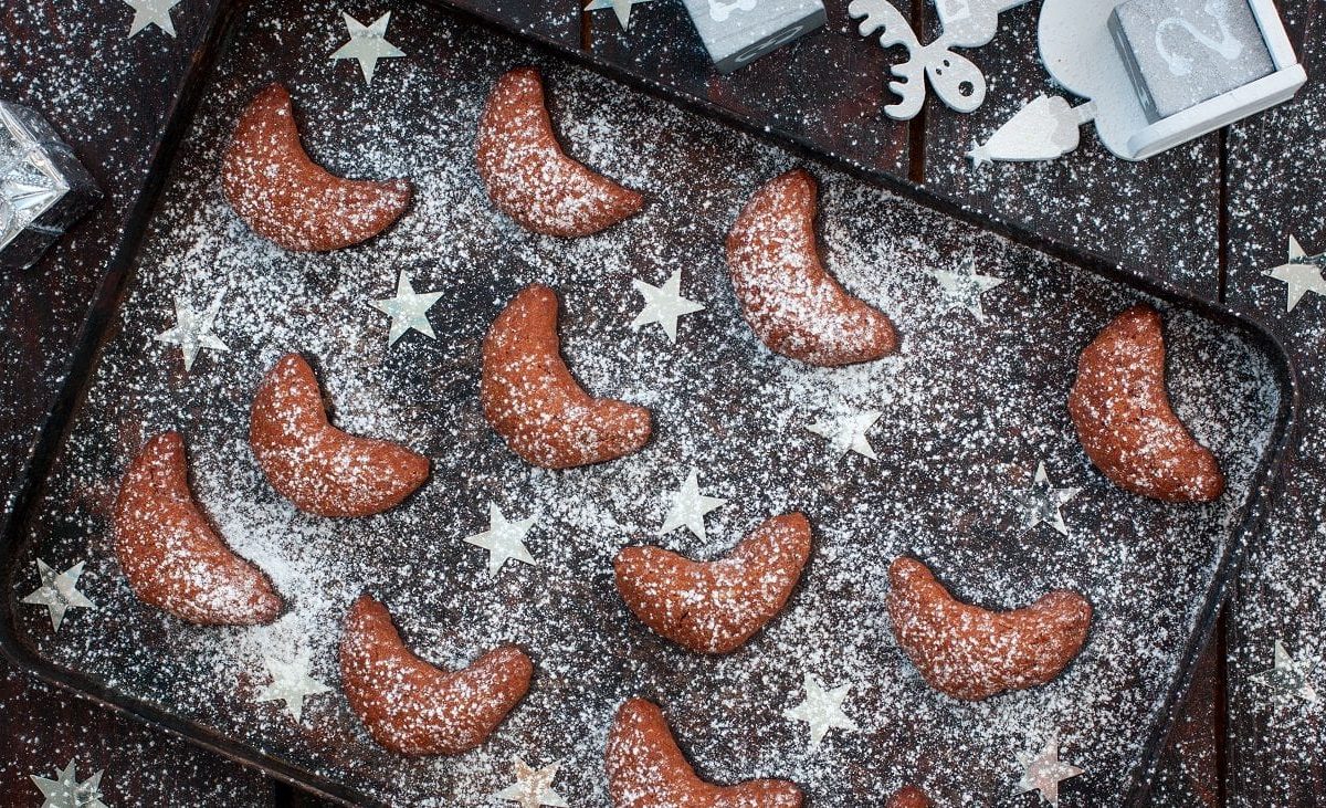 Draufsicht: Auf einem dunklen Backblech liegen dunkle schokoladen-kipferl. Die sind mit einem hellen Puder bestäubt. Dazwischen liegen Sterne aus hellem Metall.