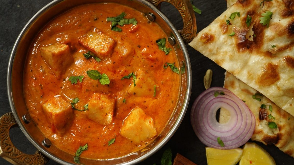 Selbst gemachter Paneer in Tomatencurry neben Zwiebel und Brot vor dunklem Hintergrund.