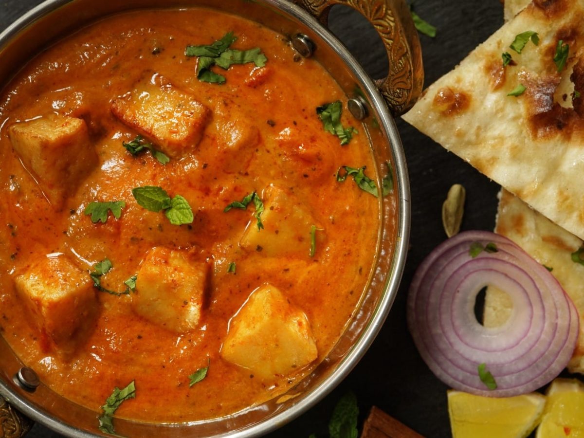 Selbst gemachter Paneer in Tomatencurry neben Zwiebel und Brot vor dunklem Hintergrund.