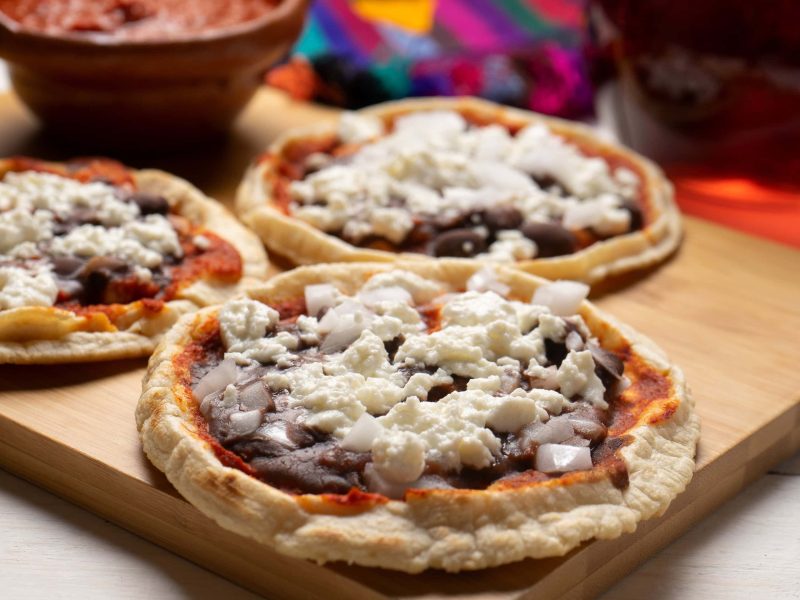 Mexikanisches Streetfood Sopes mit Bohnenmus und Käse auf Holzbrett auf Holztisch. Im Hintergrund eine Tonschale mit roter Sauce. Draufsicht.