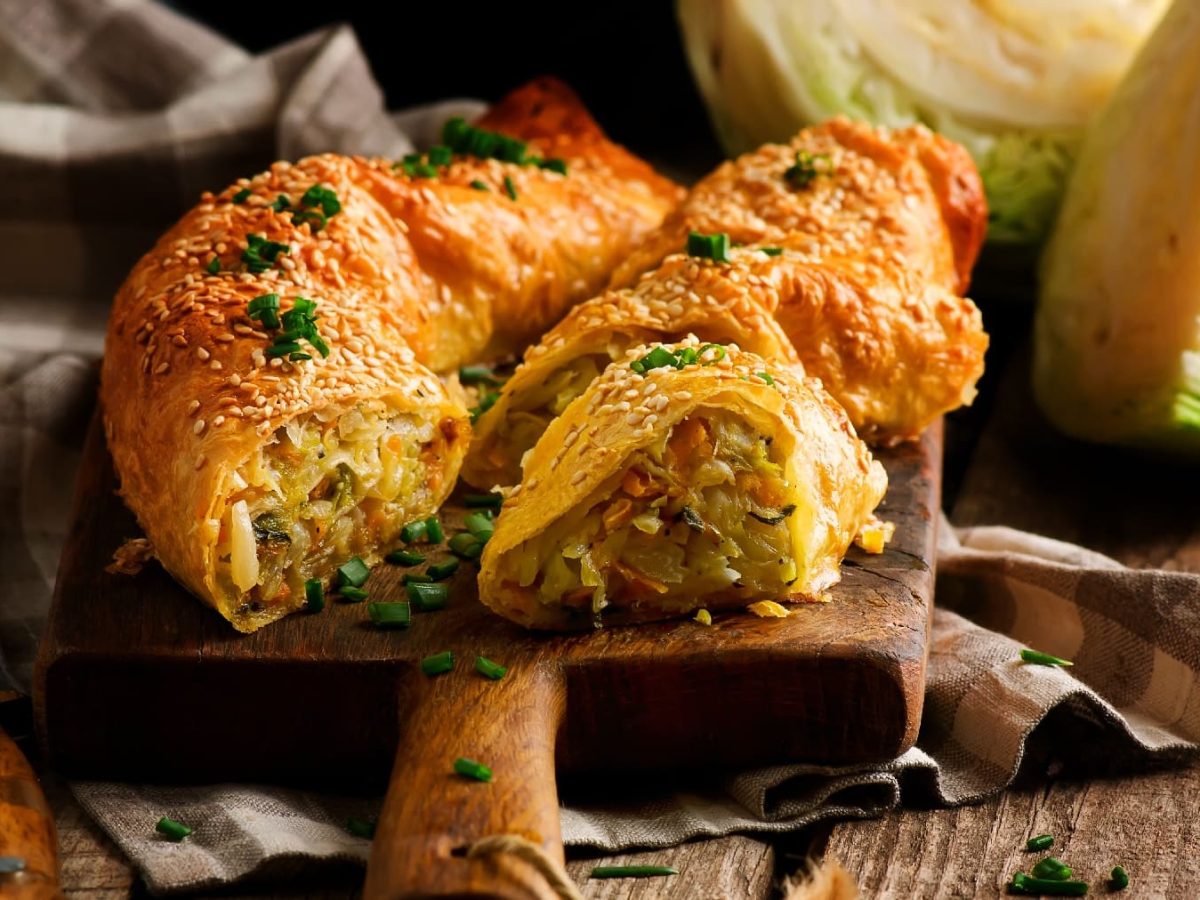 Ein Schneidebrett mit dem Strudel gefüllt mit Kohl, Kartoffeln und Karotten auf einem rustikalen Holztisch.