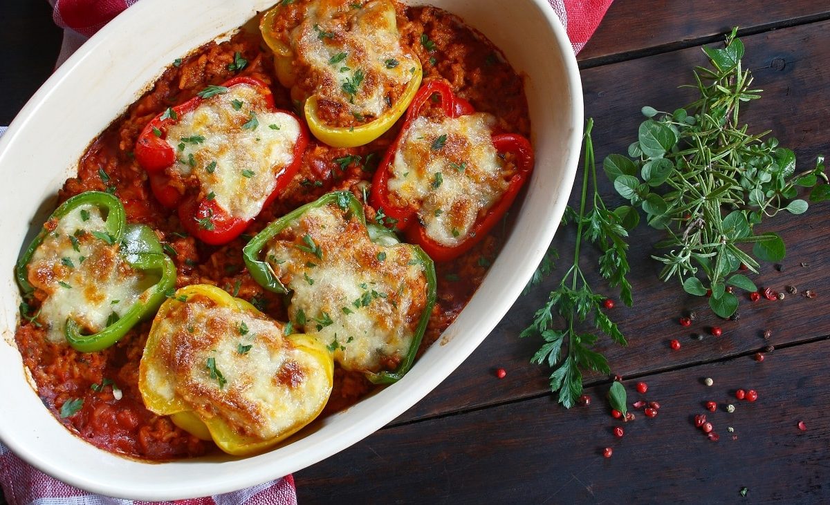 Draufsicht: Eine ofenfeste Form mit gefüllten Paprika-Hälften, die in einer Tomatensauce liegen. daneben liegen Kräuter.