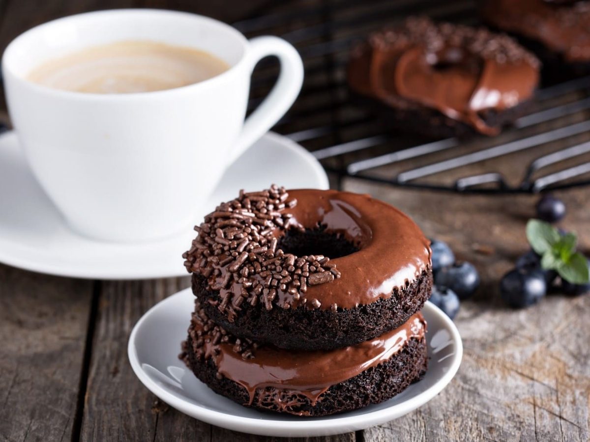 Triple Chocolate Donuts, bestehend aus Schokodunts mit Schokoglasur und Schokostreuseln, aufeinander gestapelt serviert mit einer Tasse Kaffee auf dunklem Holztisch.