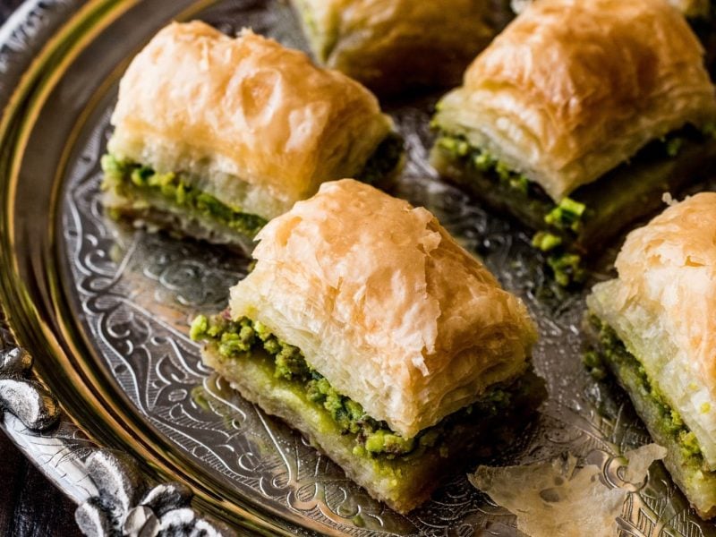 Baklava mit Pistazienfüllung auf einem silbernen Tablett vor dunklem Hintergrund.