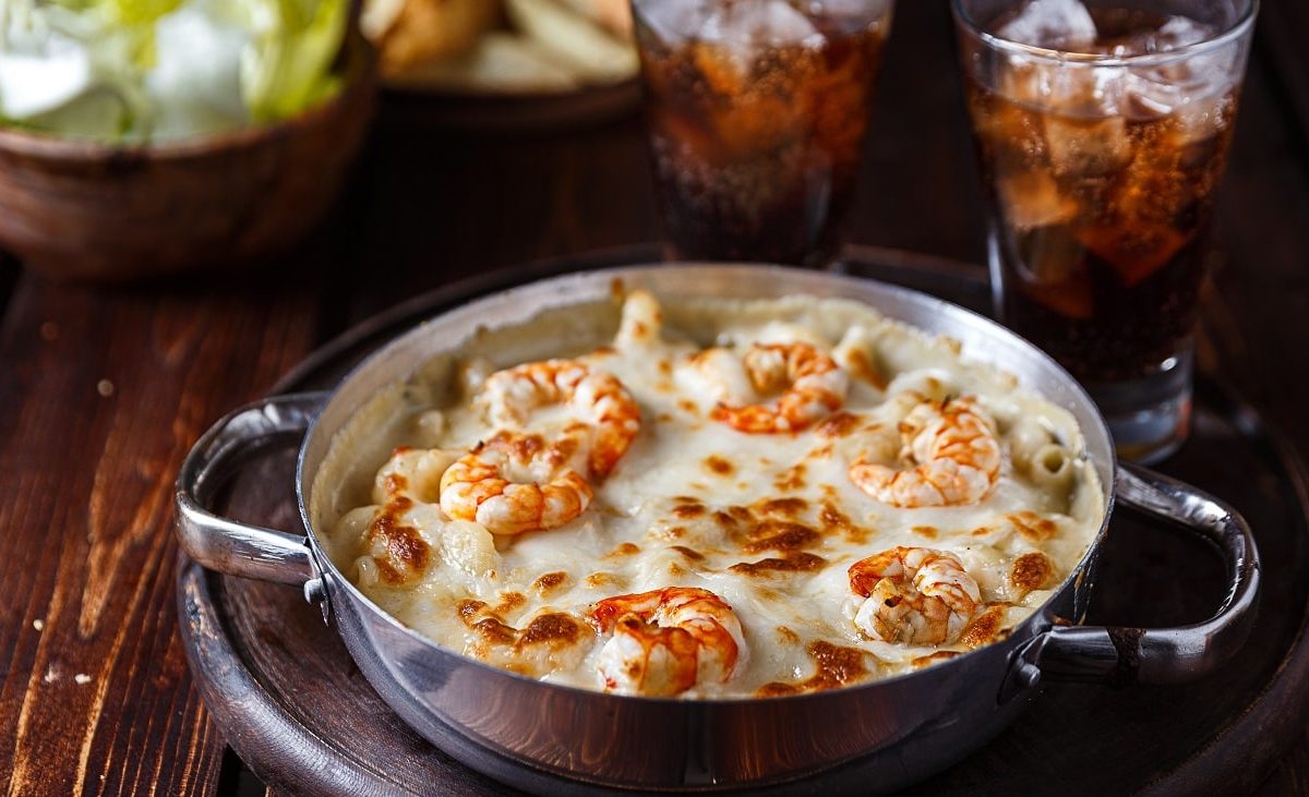 Schräge Ansicht von oben: eine dunkle Pfanne mit Überbackener Pasta mit Shrimps steht auf einem dunklen Untergrund aus Holz. Dahinter stehen zwei Gläser mit einer dunklen Flüssigkeit und Eiswürfeln.