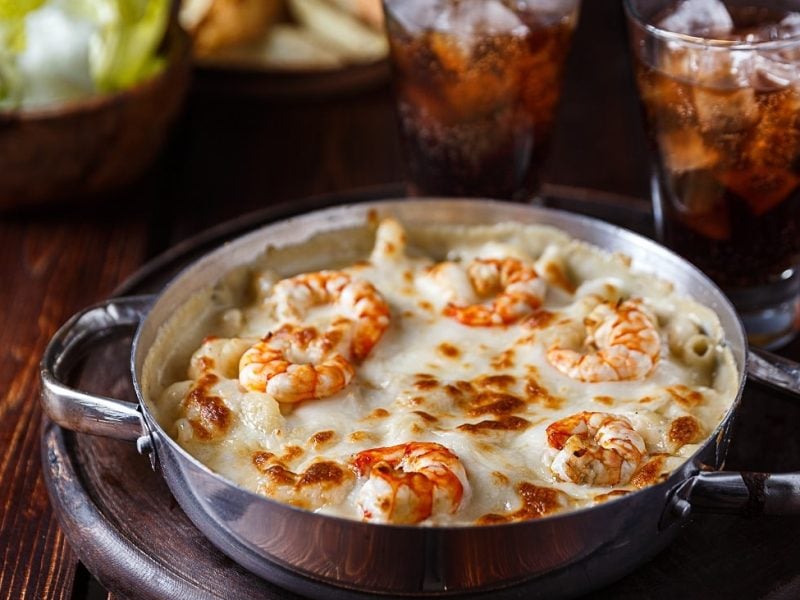 Schräge Ansicht von oben: eine dunkle Pfanne mit Überbackener Pasta mit Shrimps steht auf einem dunklen Untergrund aus Holz. Dahinter stehen zwei Gläser mit einer dunklen Flüssigkeit und Eiswürfeln.