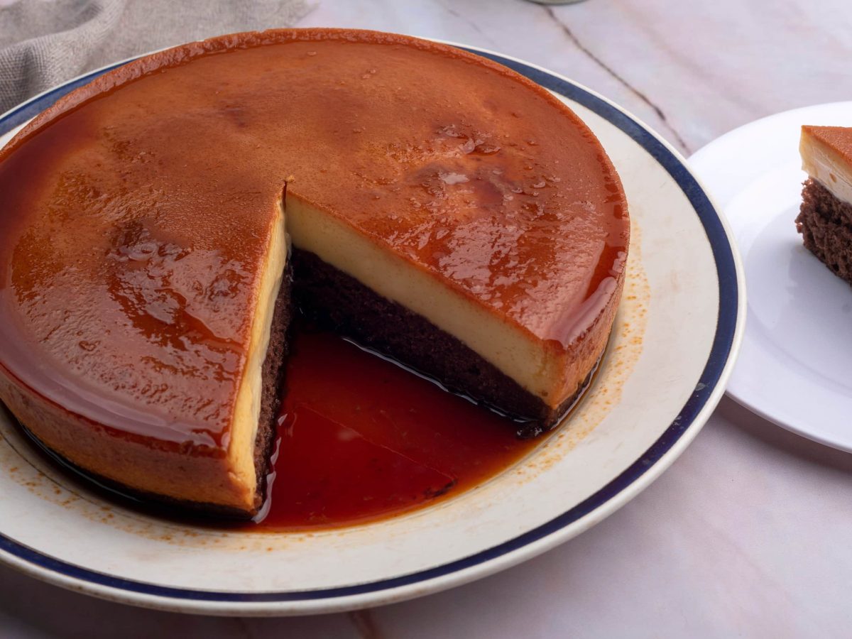 Umgedrehter Schokoladenkuchen mit Milchmädchen-Flan und Karamell auf weißem Teller auf weißem Untergrund. Daneben ein Stück Kuchen auf Kuchenteller. Draufsicht.