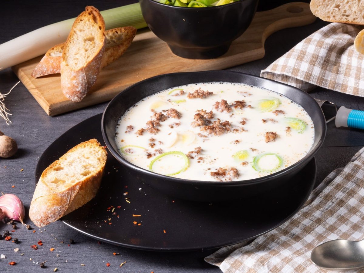 Ein tiefer Teller vegane Käse-Lauch-Suppe mit Hack, daneben Baguette, Knoblauch, Muskatnüsse und Lauch.