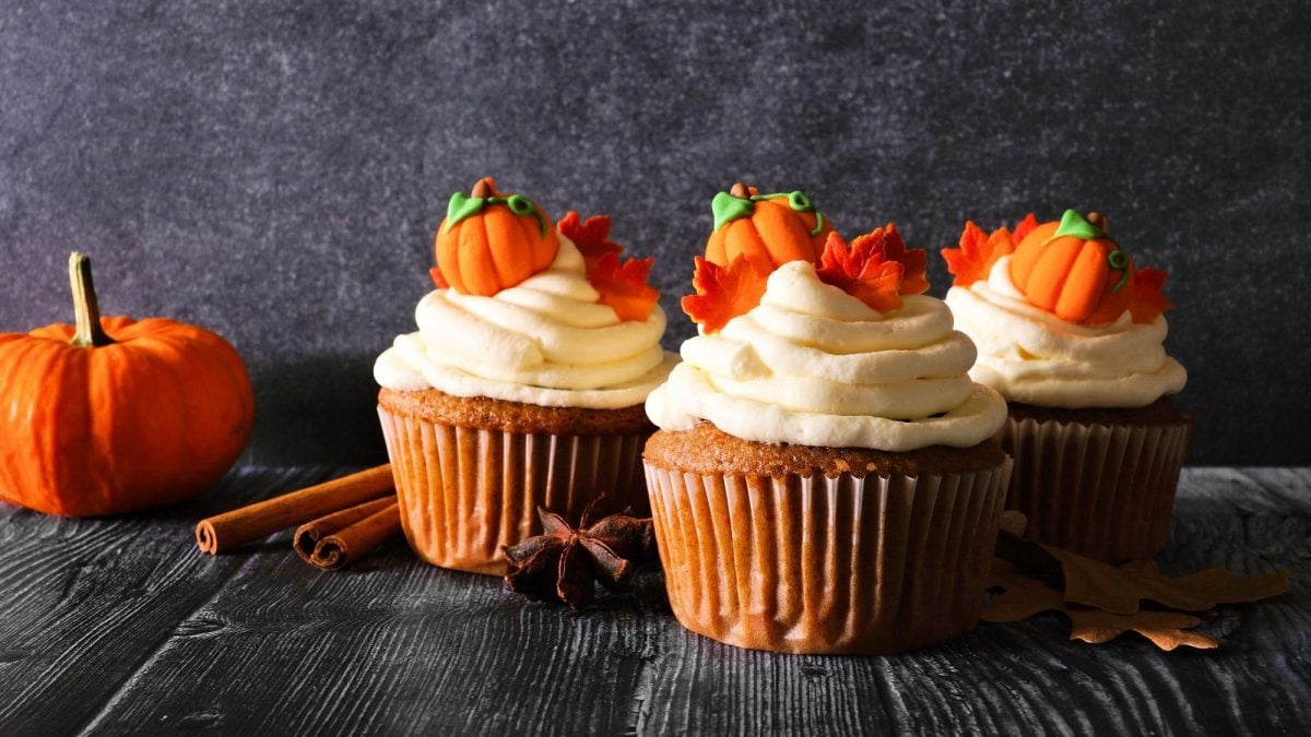 Drei Kürbis-Cupcakes mit kleinen Zuckerkürbissen verziert, daneben ein Hokkaido und Zimtstangen.