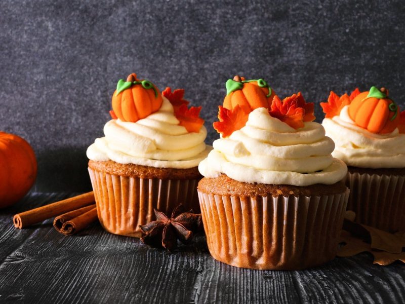 Drei Kürbis-Cupcakes mit kleinen Zuckerkürbissen verziert, daneben ein Hokkaido und Zimtstangen.