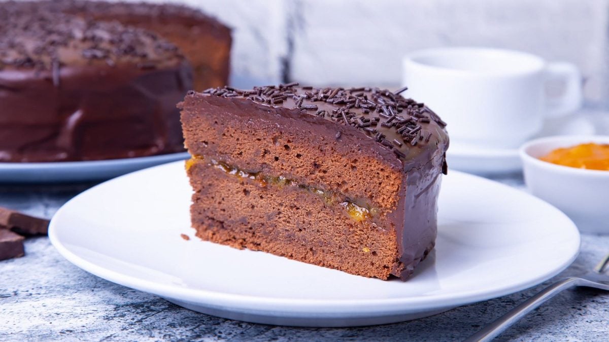 Ein Stück vegane Sachertorte auf einem Teller, im Hintergrund der angeschnittene Kuchen.