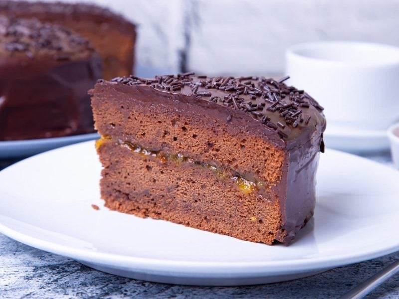 Ein Stück vegane Sachertorte auf einem Teller, im Hintergrund der angeschnittene Kuchen.