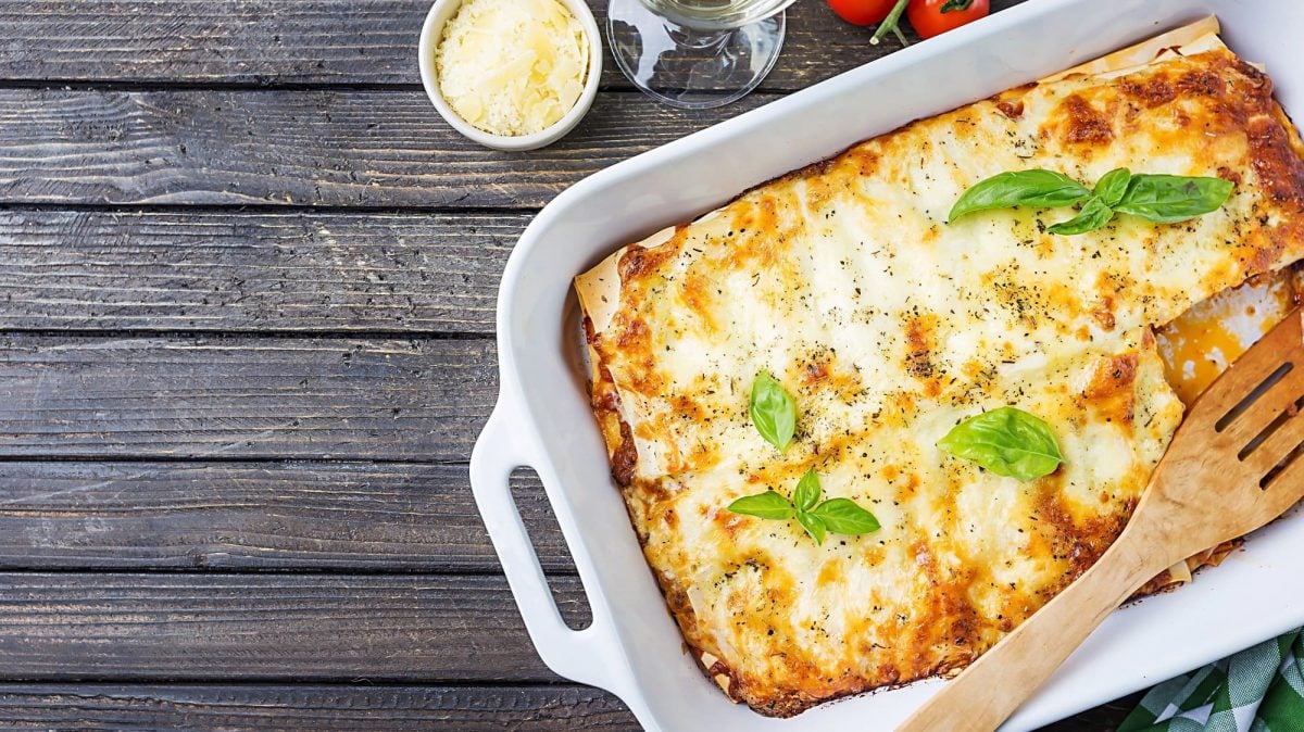 EIne Auflaufform mit veganer Spitzkohl-Lasagne und Basilikum, einem Pfannenwender, alles Draufsicht.