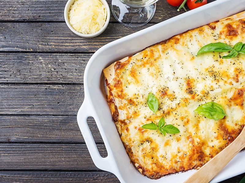 EIne Auflaufform mit veganer Spitzkohl-Lasagne und Basilikum, einem Pfannenwender, alles Draufsicht.