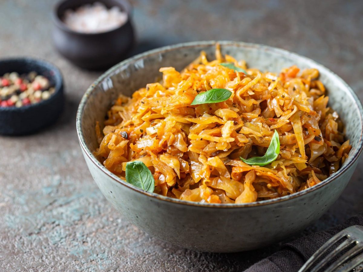 Eine Schüssel veganer Schmorkohl mit Basilikum, daneben Gabeln und Schälchen mit Gewürzen.