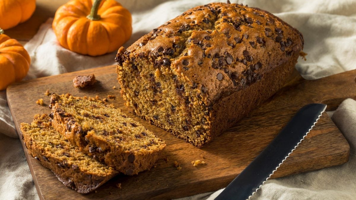 Ein angeschnittenes veganes Kürbisbrot auf einem Holzbrett, daneben Zierkürbisse und ein Brotmesser.