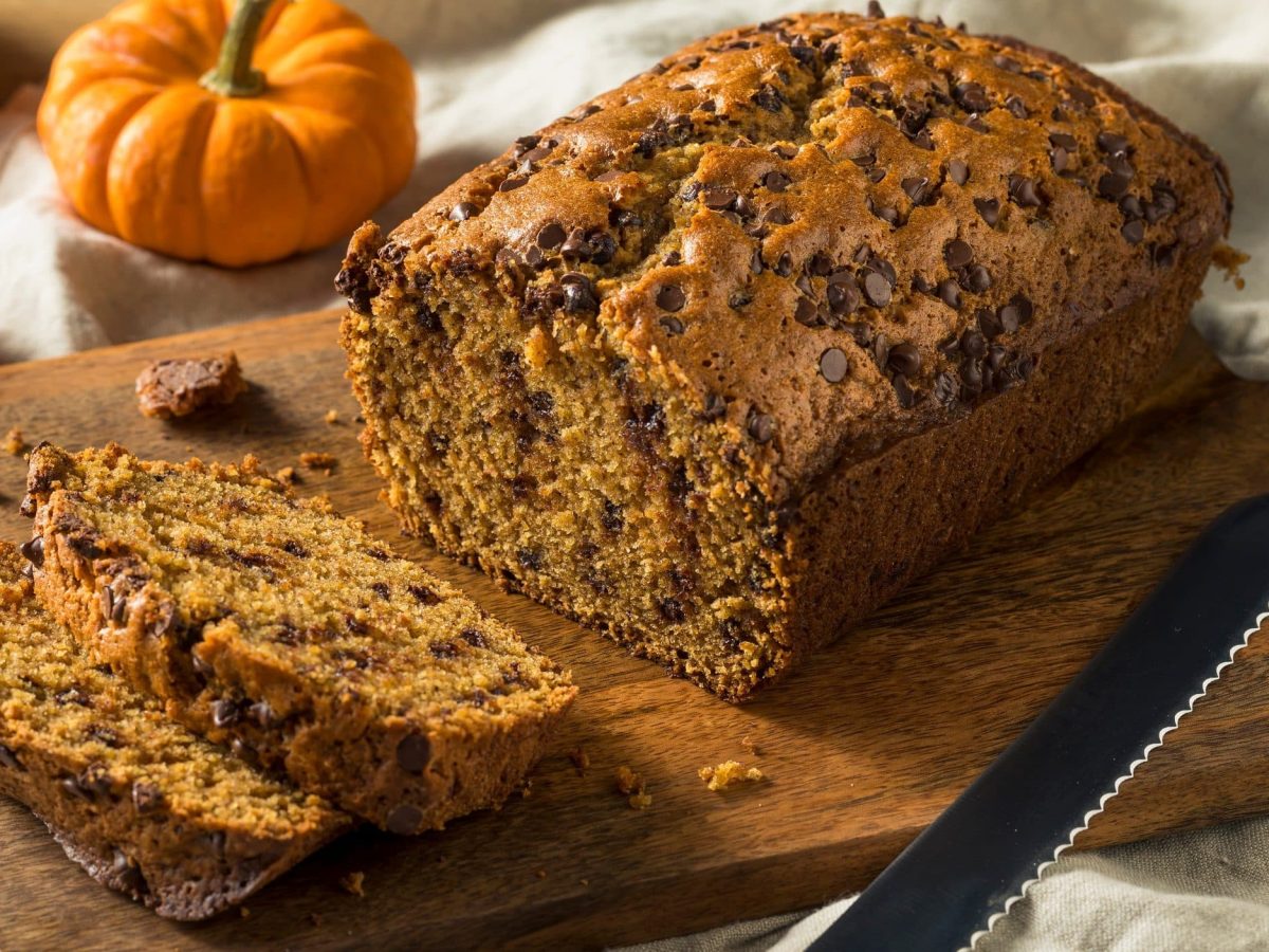 Ein angeschnittenes veganes Kürbisbrot auf einem Holzbrett, daneben Zierkürbisse und ein Brotmesser.