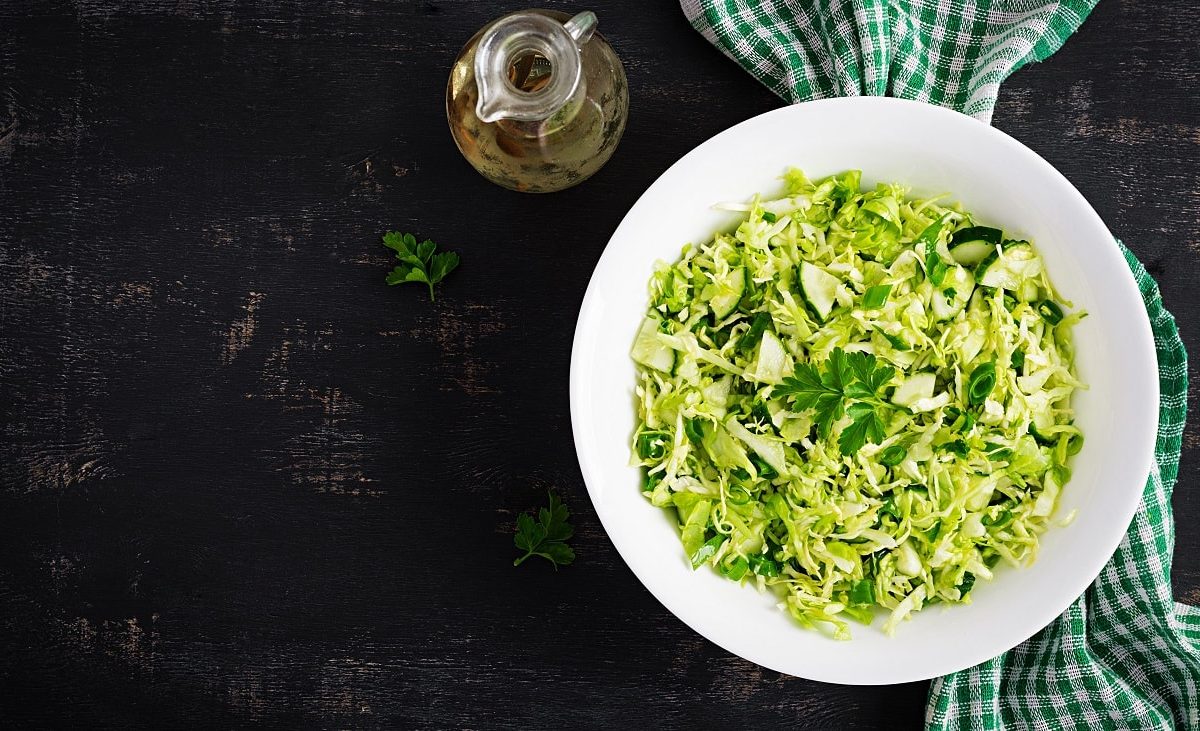 Draufsicht: Ein Salat mit Kraut, Gurke und Petersilie auf einem weißen teller steht auf einem dunklen Untergrund. Daneben liegt ein handtuch.