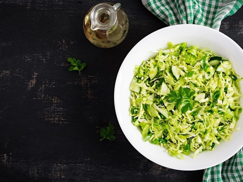 Draufsicht: Ein Salat mit Kraut, Gurke und Petersilie auf einem weißen teller steht auf einem dunklen Untergrund. Daneben liegt ein handtuch.