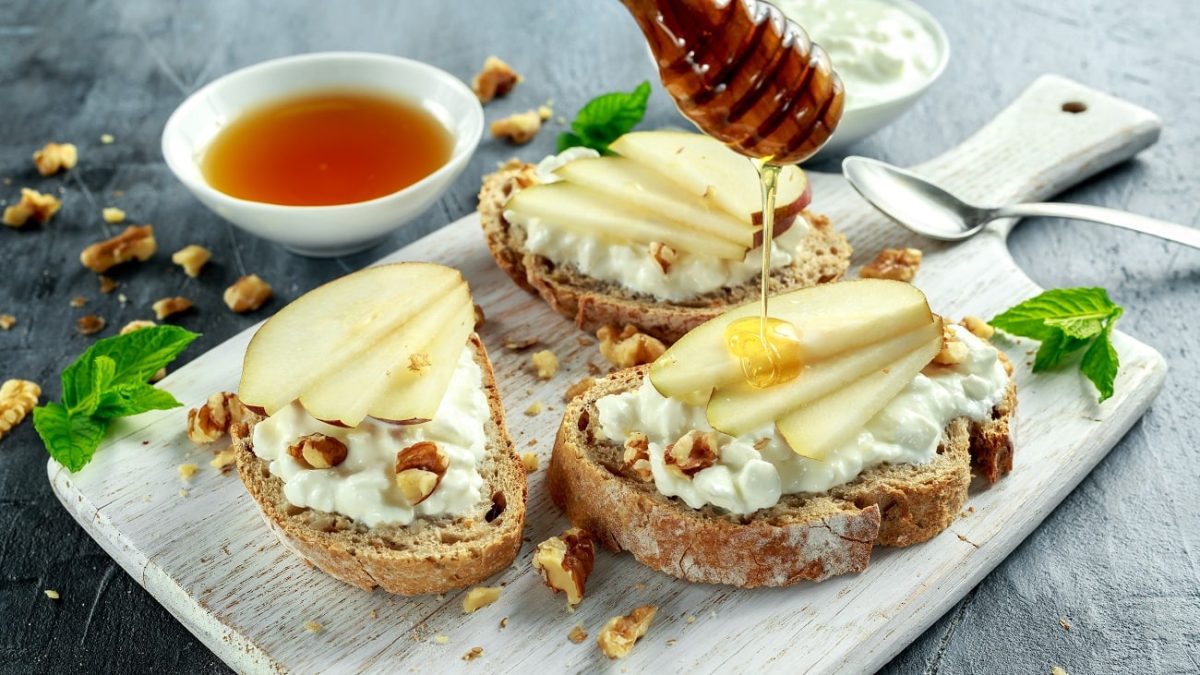 Frontalsicht: Auf einem Holzbrettchen liegen drei Ziegenkäse-Bruschetta mit Birne, Walnüssen und Honig. daneben lieben vertreut Walnusskernstücken und eine Schale mit einer dunklen Flüssigkeit.