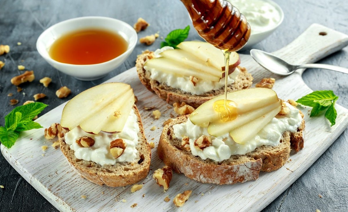 Frontalsicht: Auf einem Holzbrettchen liegen drei Ziegenkäse-Bruschetta mit Birne, Walnüssen und Honig. daneben lieben vertreut Walnusskernstücken und eine Schale mit einer dunklen Flüssigkeit.