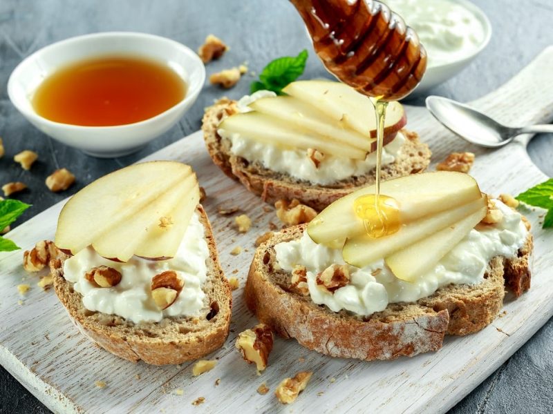 Frontalsicht: Auf einem Holzbrettchen liegen drei Ziegenkäse-Bruschetta mit Birne, Walnüssen und Honig. daneben lieben vertreut Walnusskernstücken und eine Schale mit einer dunklen Flüssigkeit.