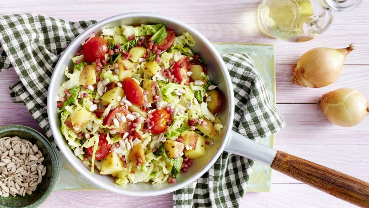 Kartoffel-Wirsing-Pfanne mit Tomaten, Speck und Sonnenblumenkernen, serviert in einer großen Pfanne auf einem Küchentuch.