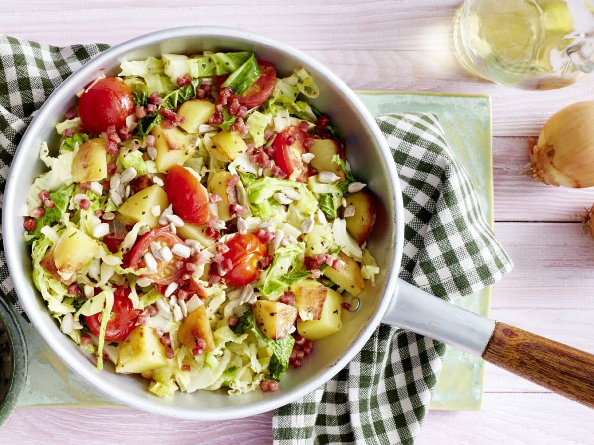 Kartoffel-Wirsing-Pfanne mit Tomaten, Speck und Sonnenblumenkernen, serviert in einer großen Pfanne auf einem Küchentuch.