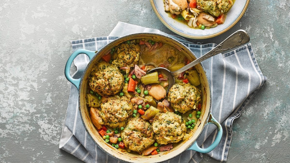 Hähnchen-Schinken-Auflauf mit Senf-Knödeln, Gemüse und frischer Petersilie, serviert in einem großen Topf auf grauem Untergrund.