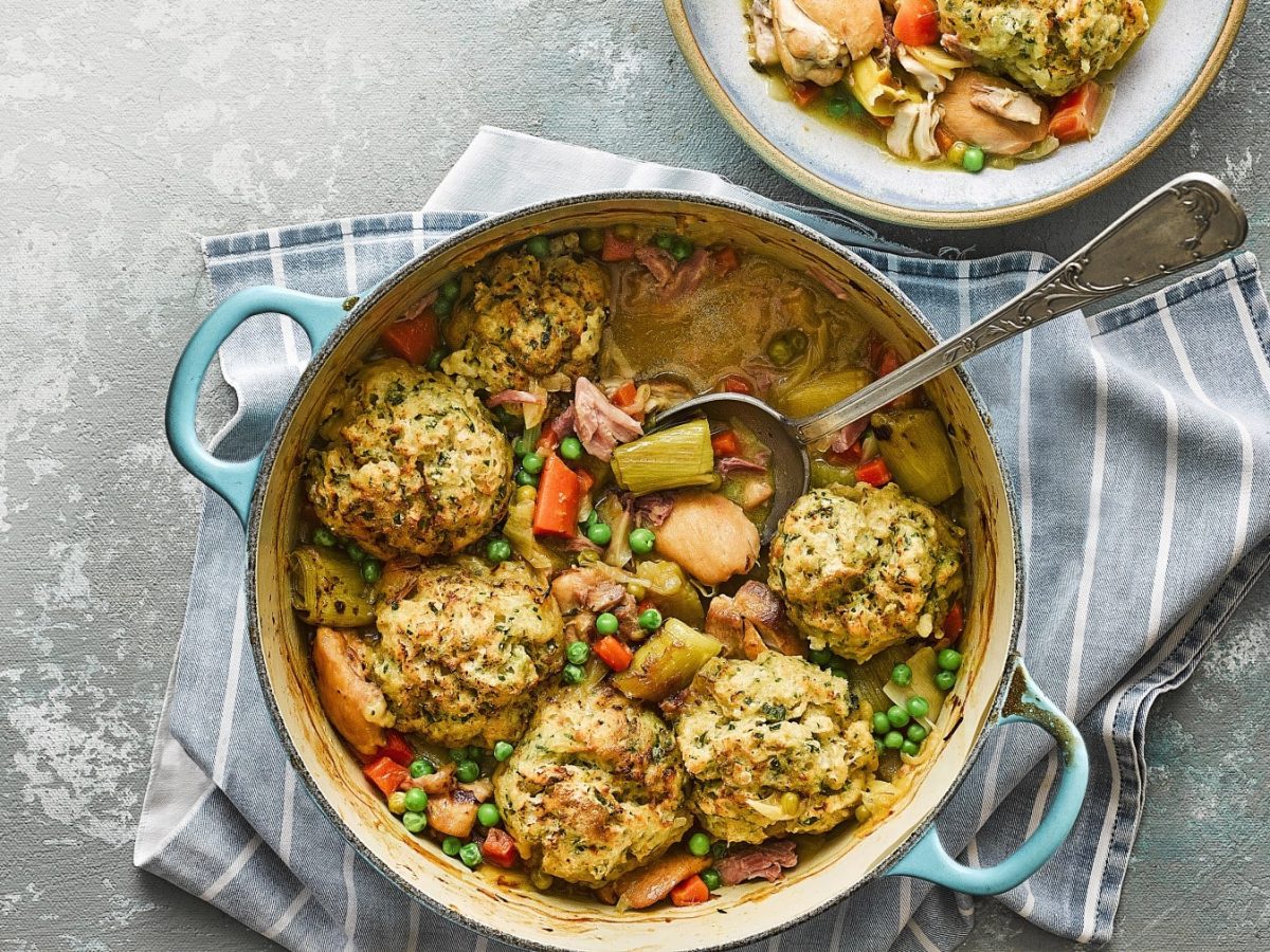 Hähnchen-Schinken-Auflauf mit Senf-Knödeln, Gemüse und frischer Petersilie, serviert in einem großen Topf auf grauem Untergrund.